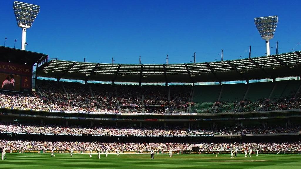The green grass of the MCG with players wearing white test kit and the stands with the crowd in the background
