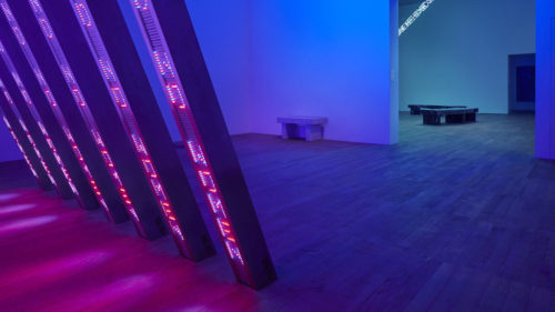 Jenny Holzer Blue Purple Tilt Tate room with Artwork from Jenny Holzer. The artwork is composed by a group of metallic bars with text displayed on their surface. The text is made of bright purple led