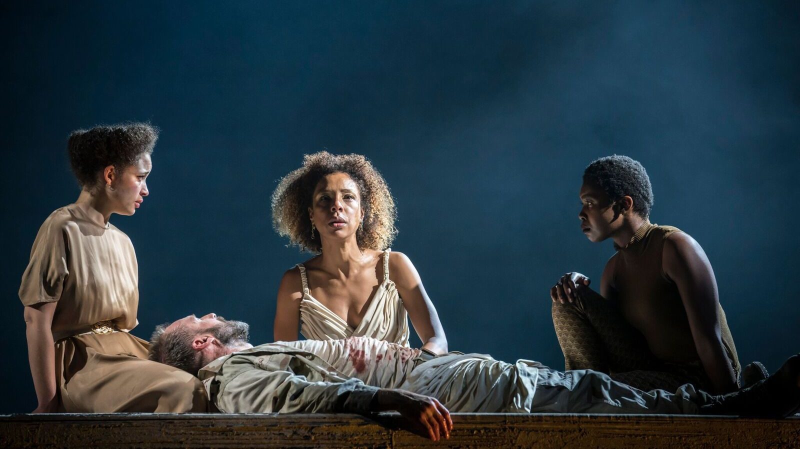 Cleopatra (Sophie Okonedo) kneels behind a dying Antony (Ralph Fiennes) flanked by two servants.