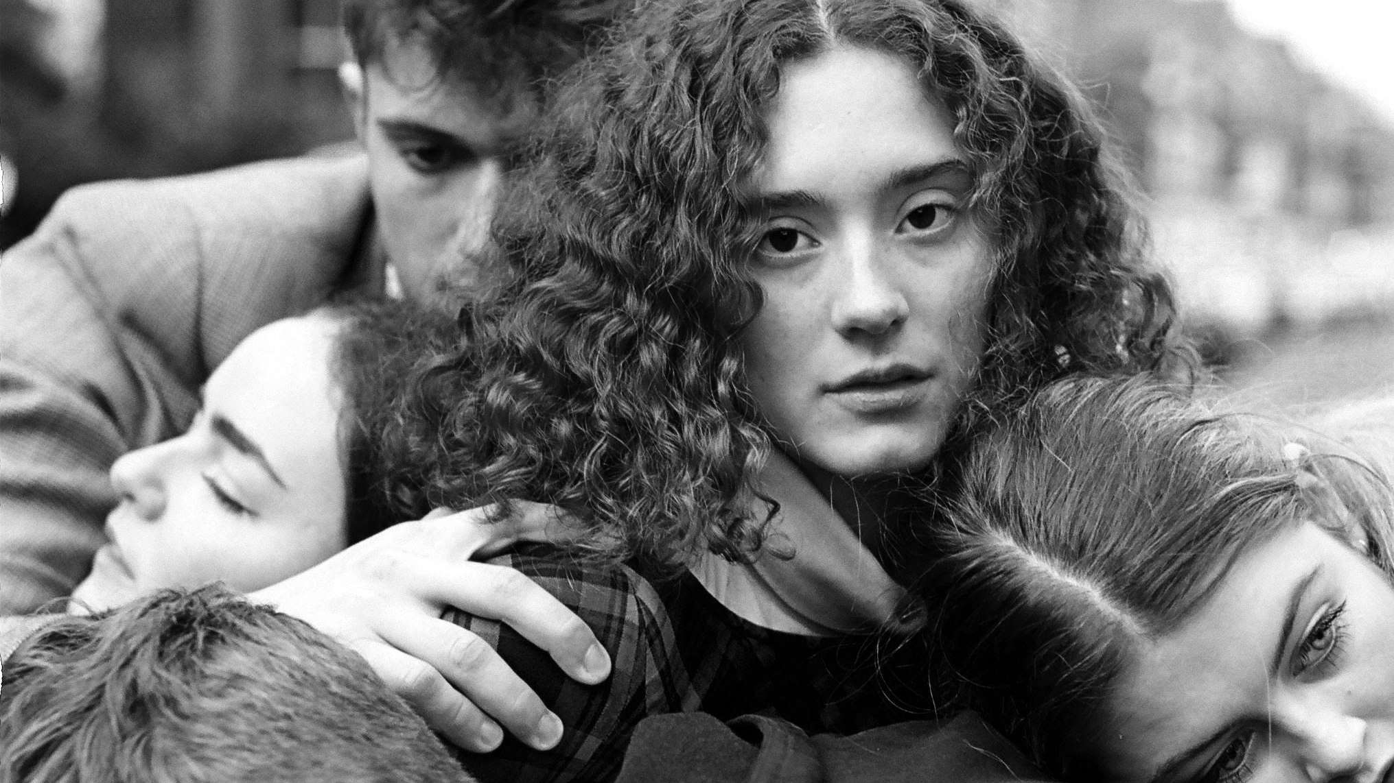 A cluster of people wrapped around one another in a greyscale photo. The central woman looks directly at the camera.