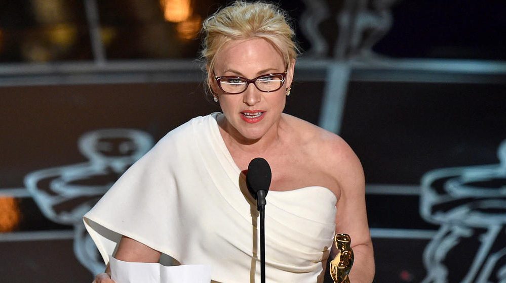 Patricia Arquette stands on stage with an Oscar statuette and a sheet of paper, speaking into a microphone.