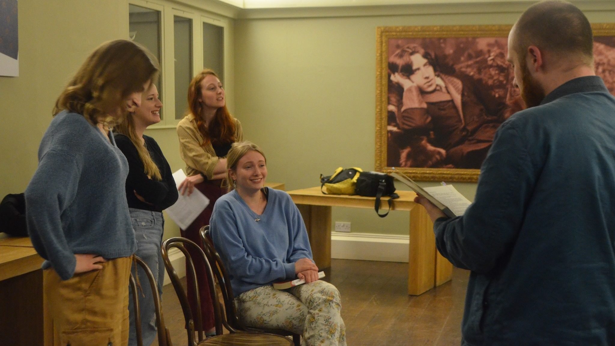 A group of four young women and a young man rehearsing a scene from a play. There is a picture of Oscar Wilde on a wall in the background