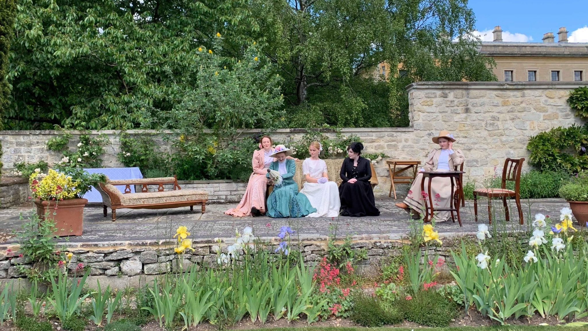 five woman dressed in old-fashioned upper class dresses sitting in a garden setting; there is a chaise long, a sofa and a table with chairs
