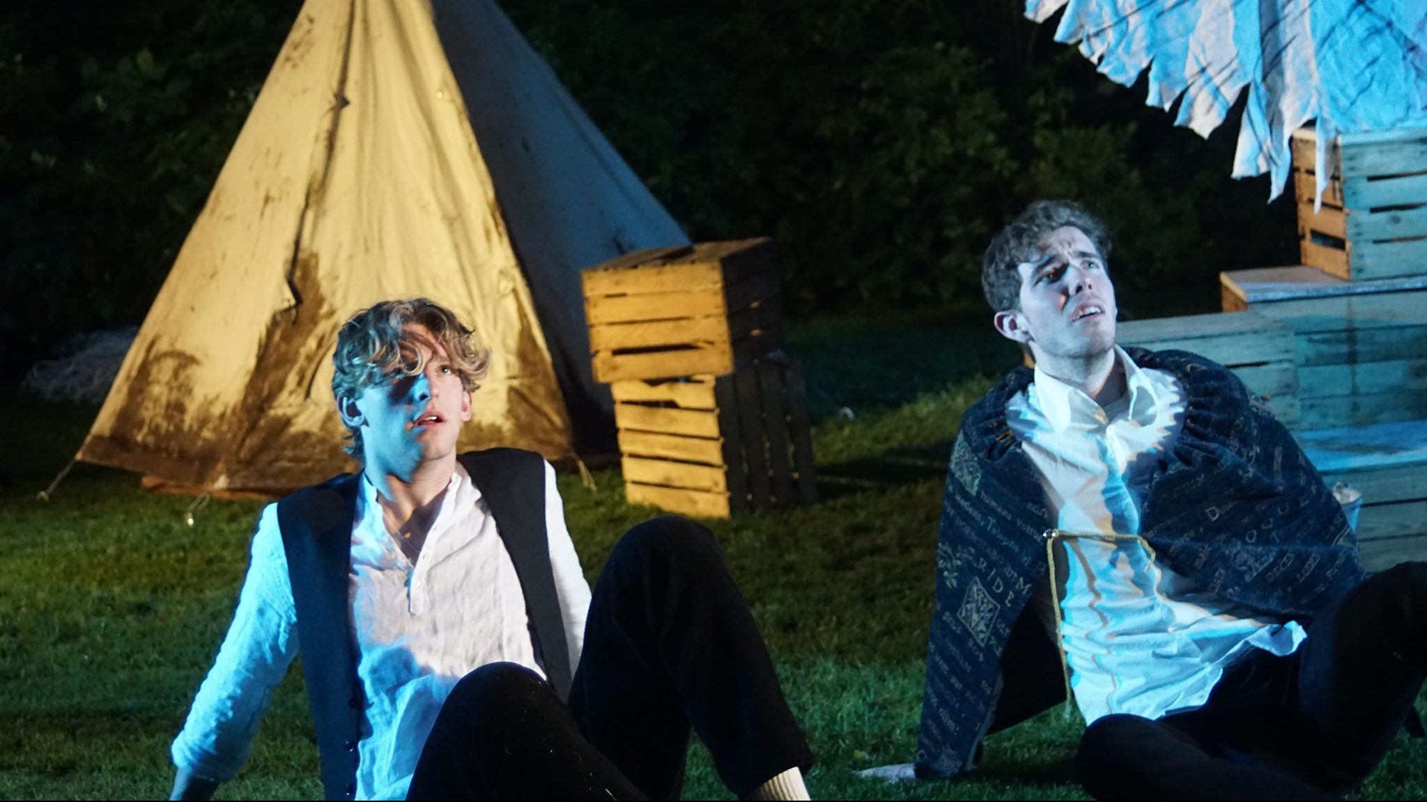 Two men sit on the floor in a blue light looking up with a tent behind them