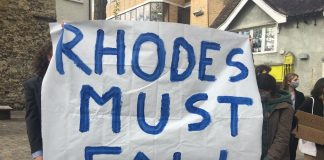 A white banner being held up by two people who one are hidden behind it. In blue letters is written: "RHODES MUST FALL"