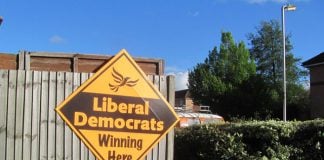 A diamond-shaped yellow sign saying "Liberal Democrats Winning Here" attached to a wooden fence.