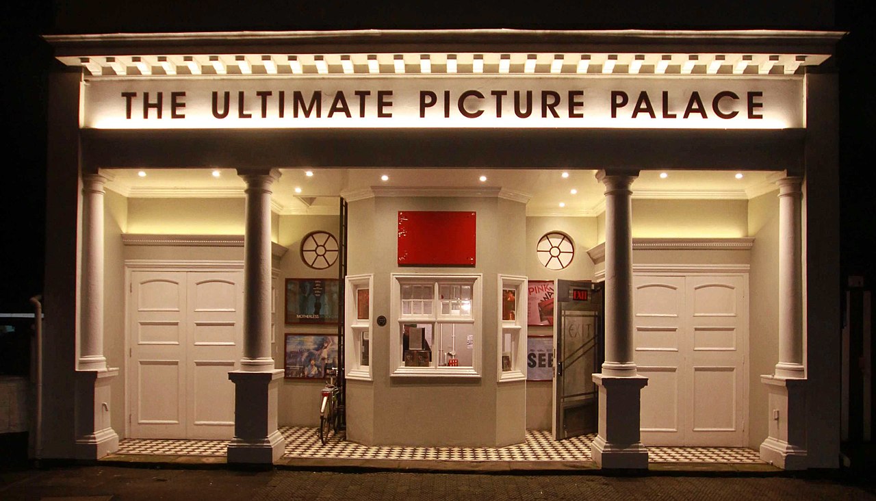 A cinema front, with the name written at the top is THE ULTIMATE PICTURE PALACE. There are two then pillars at the front, at between these two pillars in the ticket office. The cinema front is lit up.