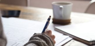 A close-up view of a desk with paper and a mug on it, and a person's arm holding a pen and making notes.