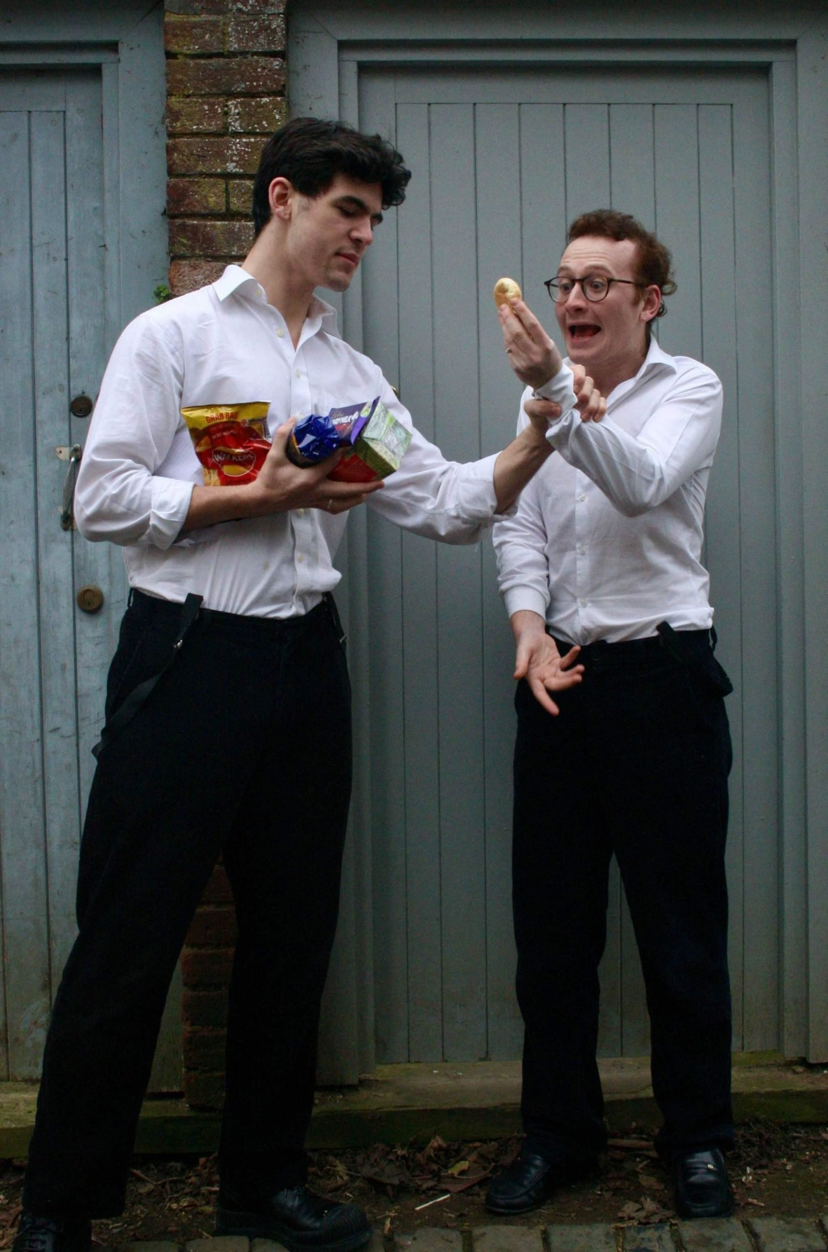 A man in a white shirt holds an armful of snacks and grabs the wrist of another man in a white shirt, who is holding an Edcles cake and looking scared.