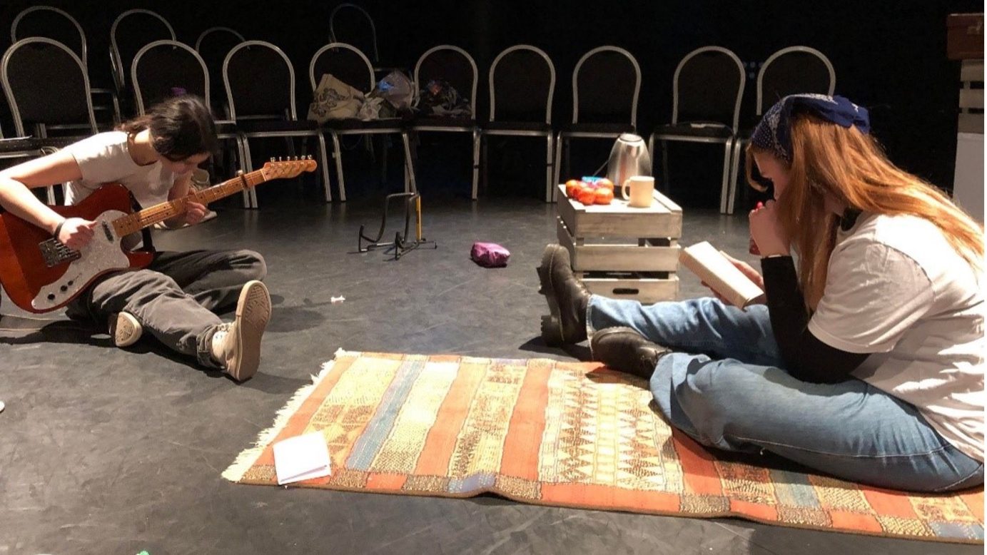 One cast member sits on the left playing the guitar, whilst the other sits on the right side of the set, reading a book.