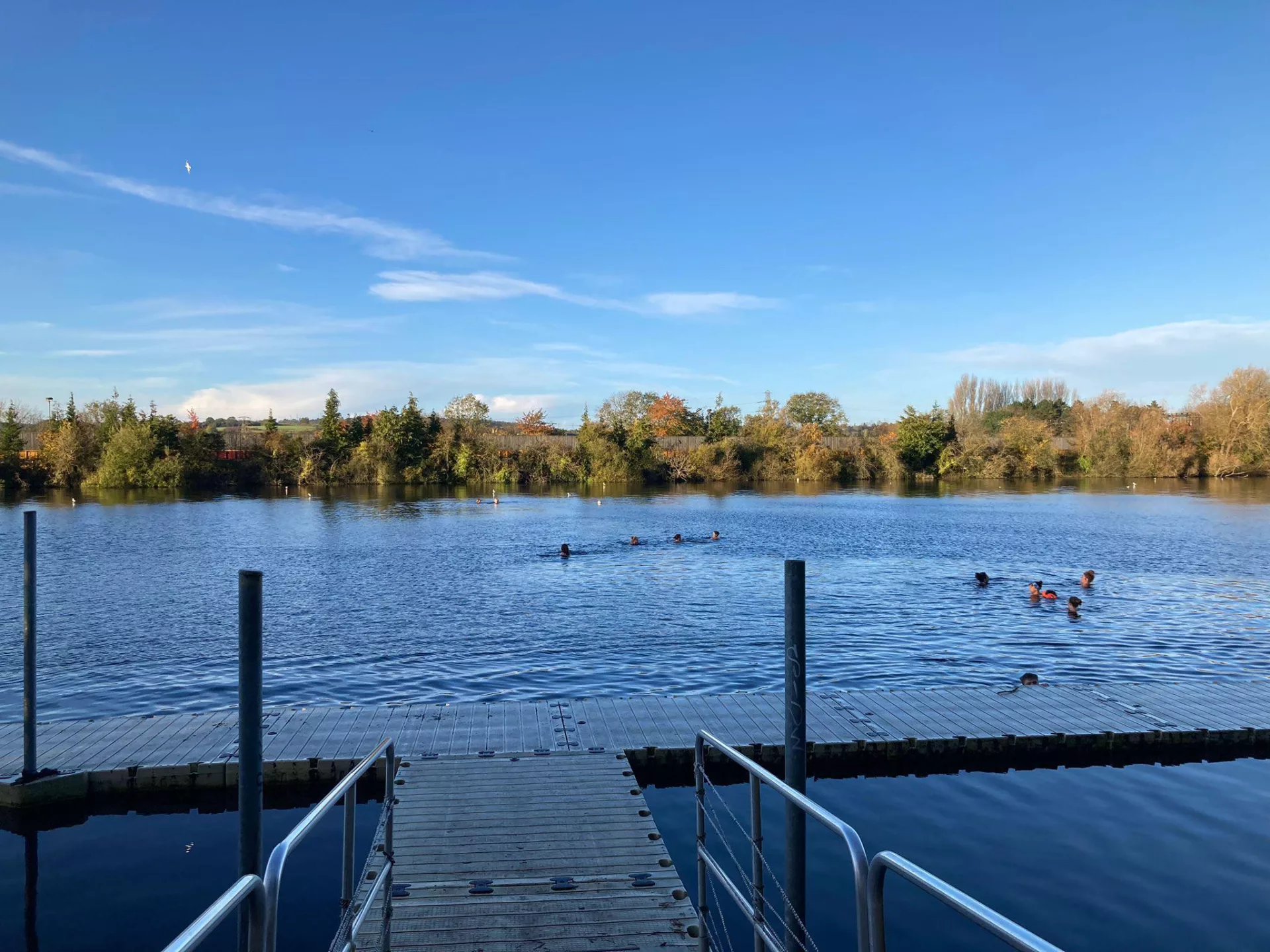 Hinksey lake looking tranquil and blue with a few swimmers and a jetty in the foreground.